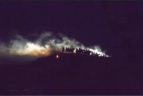 A group of horsemen silhouetted against a ridge at night with smoke and light behind them.