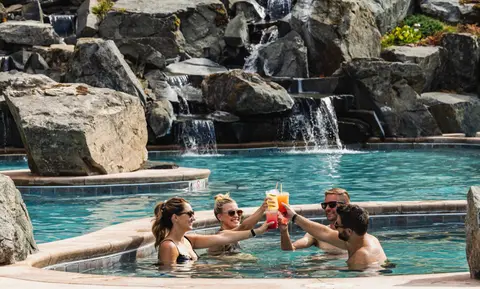 Four people toast with drinks in a resort pool in front of a rock waterfall.