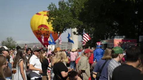 A crowd of people gathers outdoors near a hot air balloon and flags.