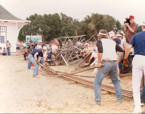 People lifting and moving metal rails near a building and a crowd.