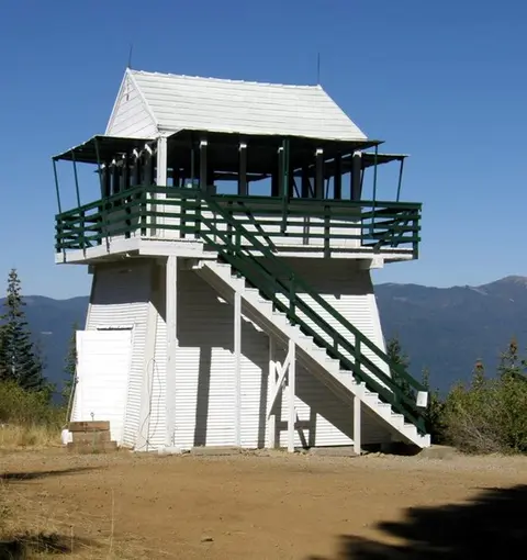 A white and green fire lookout tower stands on a mountaintop against a clear blue sky.