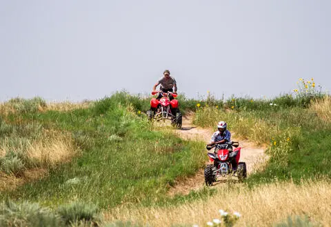 Two people ride all-terrain vehicles along a dirt path through grassy hills.