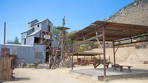 An outdoor historic industrial site with a corrugated metal mill building, wooden structures, and a desert landscape.