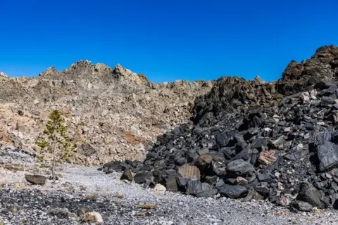 A rugged landscape features piles of dark and light rocks with a small tree under a blue sky.