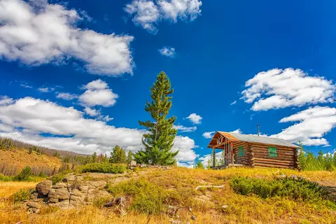 A rustic log cabin sits on a grassy hill under a bright blue sky with clouds.