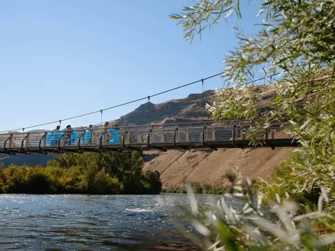 People walking across a suspension bridge with inner tubes over a river.