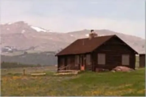 A small cabin sits in a field with mountains in the background.