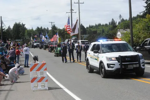 A color photograph shows a parade on a street with a police vehicle leading the way.