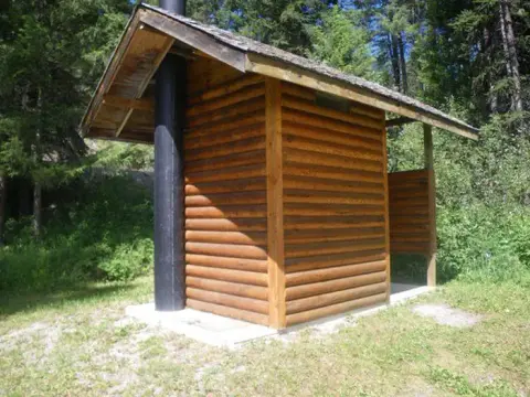 A small wooden outhouse with a black pipe stands in a forest clearing.