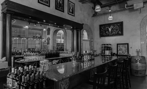 Black and white photograph showing the interior of a winery or bar with a counter, stools, and wine bottles.