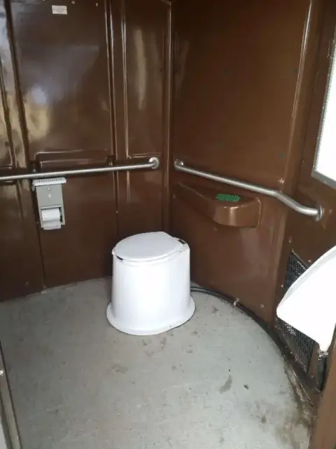 The interior of a portable toilet with a white commode and handrails.