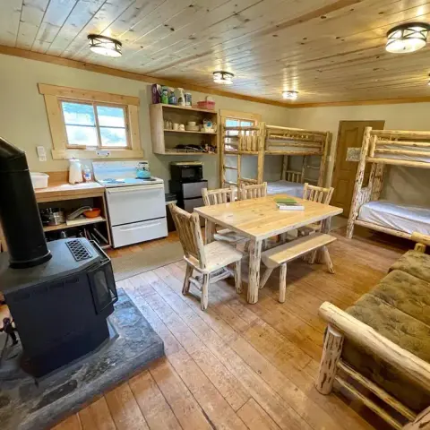 A rustic cabin interior featuring bunk beds, a wooden dining table, and a small kitchen area.