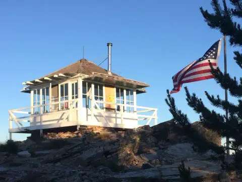 A fire lookout cabin stands on a rocky summit with an American flag flying nearby.