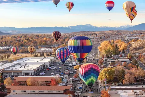 Multiple hot air balloons fly over a town with mountains in the distance.
