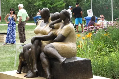 A sculpture of three figures sitting together outdoors, with people and garden flowers in the background.