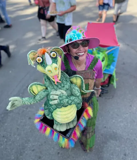 A woman smiling while operating a green dragon puppet outdoors.