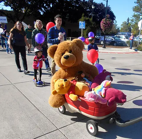 People walk down a street with a red wagon filled with stuffed animals and balloons.