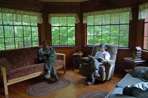 Two people read books inside a wood-paneled room with large windows.