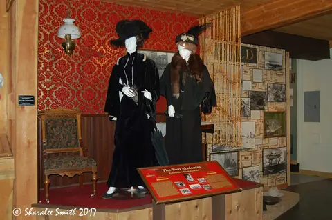 An indoor museum exhibit shows two mannequins dressed in period attire with a chair and informational sign.