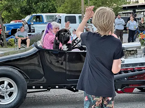 A woman and her dog wearing sunglasses ride in a custom hot rod car.