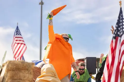 A person dressed in a carrot costume holds a large carrot in the air outdoors with American flags nearby.