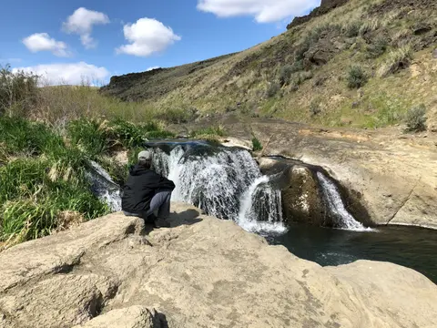 A person sits on a rock by a small waterfall in a landscape.