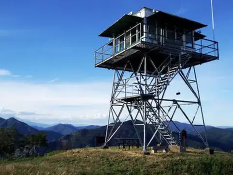 A fire lookout tower stands on a grassy mountain peak overlooking distant ranges.