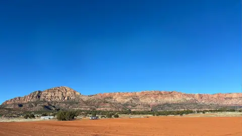 A wide shot of a desert landscape with red earth, low green vegetation, and rocky mountains under a clear blue sky.