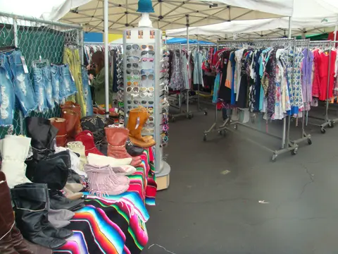 An outdoor market stall displaying rows of clothing, boots, and sunglasses under tents.