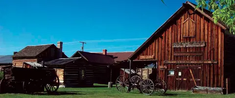 Historic wooden buildings and wagons stand in a grassy area under a clear blue sky.