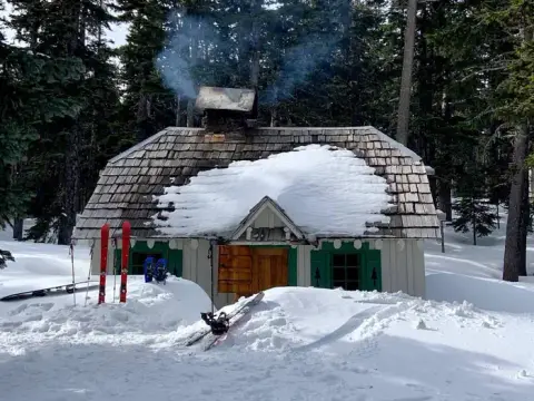 A snowy cabin in the woods with smoke coming from the chimney and skis propped outside.