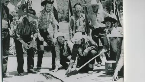 A historic black and white photograph showing a group of men gathered around a large frog on the ground.