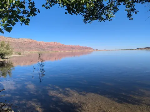 A calm lake reflects distant red rock cliffs under a clear blue sky.