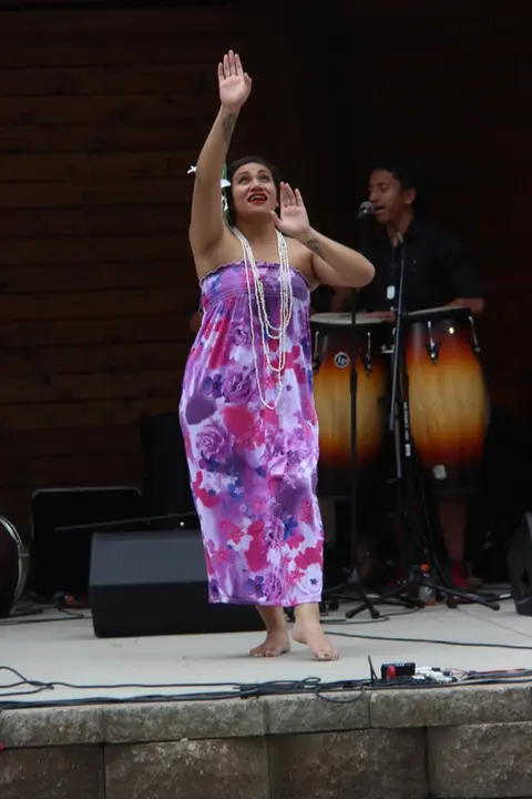 A woman in a floral dress dances on stage with a musician in the background.