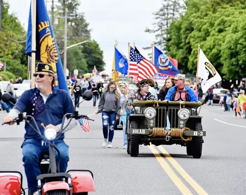 People participate in a street parade with flags and a vintage military vehicle.