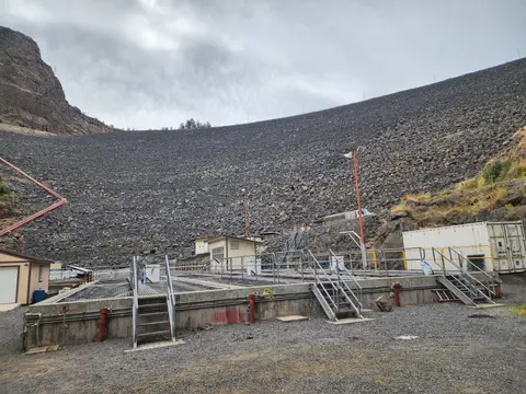 A large rock-fill dam with industrial structures in front under a cloudy sky.