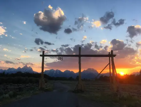 A wooden ranch gateway stands before a scenic mountain landscape under a sunset sky.