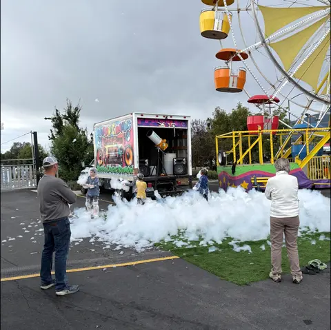 People watch children playing in a pile of foam near a carnival truck and Ferris wheel.