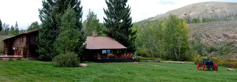 Exterior view of a rustic log cabin set in a grassy landscape with trees and hills.
