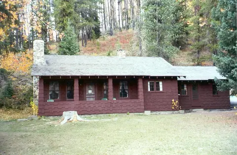 A brown wooden cabin with a stone chimney sits among trees in the forest.