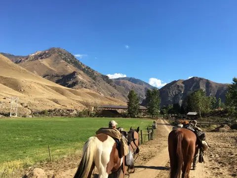 People ride horses along a dirt path near a grassy field and mountains.