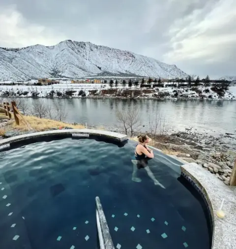 A person relaxing in an outdoor hot tub overlooking a river and snow-covered mountains.