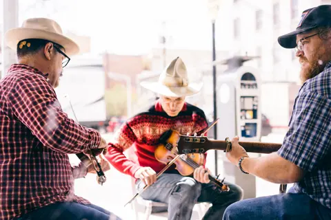 Three musicians play stringed instruments outdoors on a sunny day.
