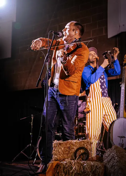 A musician plays a fiddle on stage while singing into a microphone, with another band member playing a tambourine.