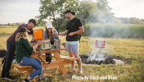 A group of friends camping and cooking by a campfire in a field.