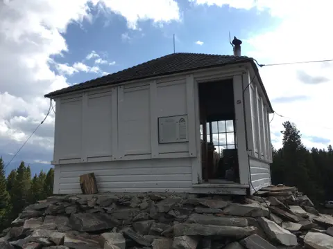 A small white wooden lookout cabin sits atop a rocky mountaintop under a cloudy blue sky.