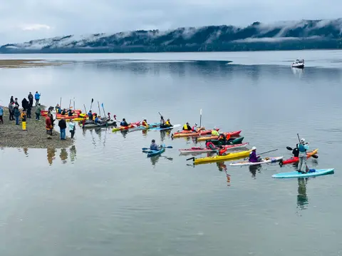 A group of people in kayaks and paddleboards gather on the water near a shore.