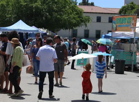 People walk around an outdoor market with booths and umbrellas.