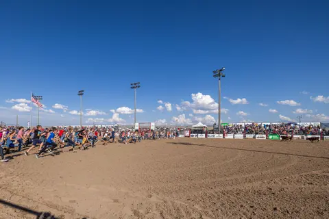 A large group of people runs across a dirt arena during an outdoor event.
