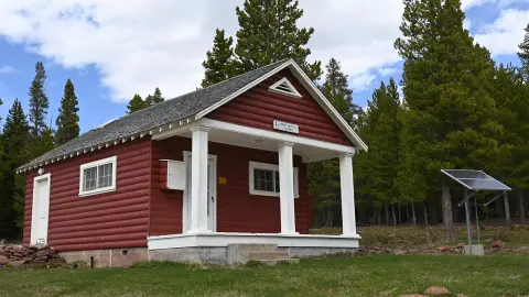 A red wooden cabin with a porch stands among pine trees.